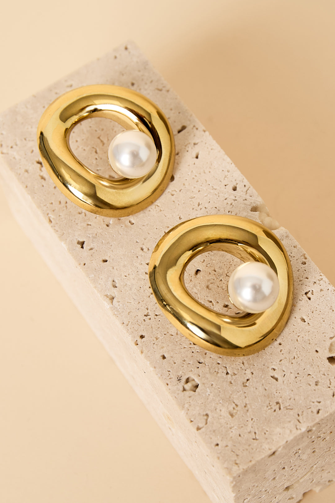 Gold circle stud earrings with a pearl accent, resting on a textured stone.