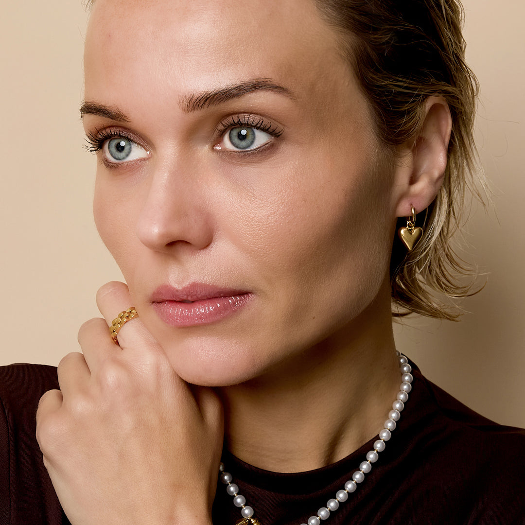 Gold heart drop earrings on a woman wearing a pearl necklace and a gold ring.