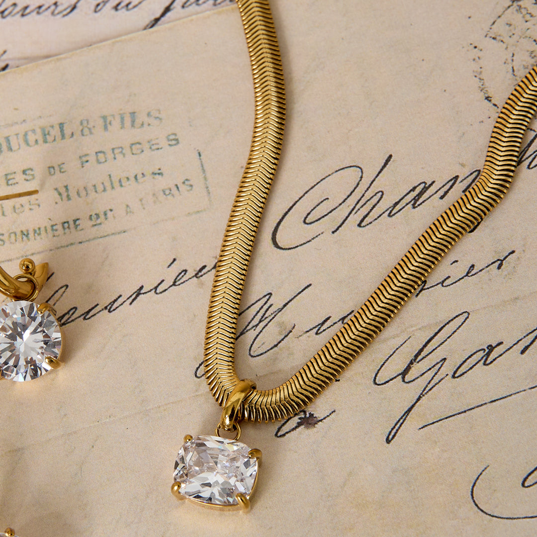 Gold snake chain necklace with a cushion-cut clear stone pendant, resting on vintage paper.