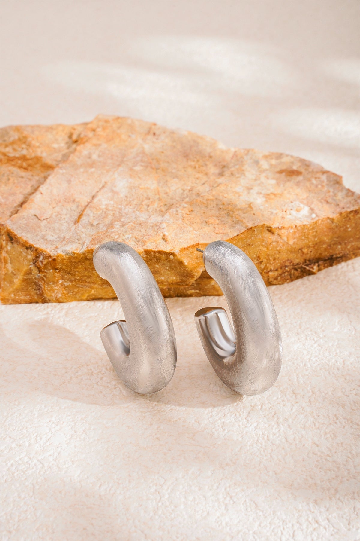 Elysian Hoop Earrings in a brushed silver finish, resting on a textured surface next to a rock.