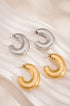 Two pairs of chunky hoop earrings, one in silver and one in gold, displayed on a white wavy surface.