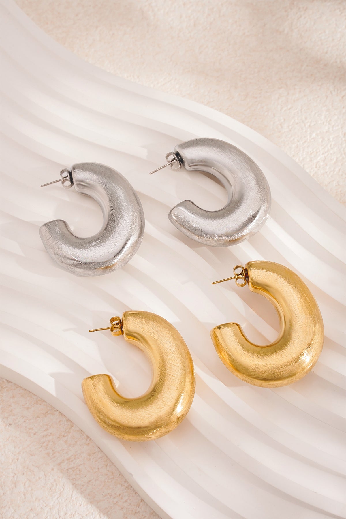 Two pairs of chunky hoop earrings, one in silver and one in gold, displayed on a white wavy surface.