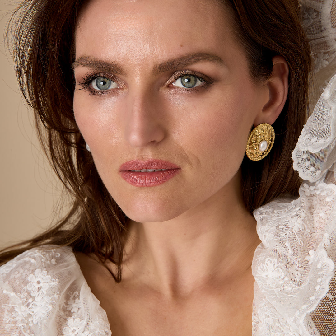 Gold Aurelia stud earrings with pearl detail worn by a woman with blue eyes and brown hair.