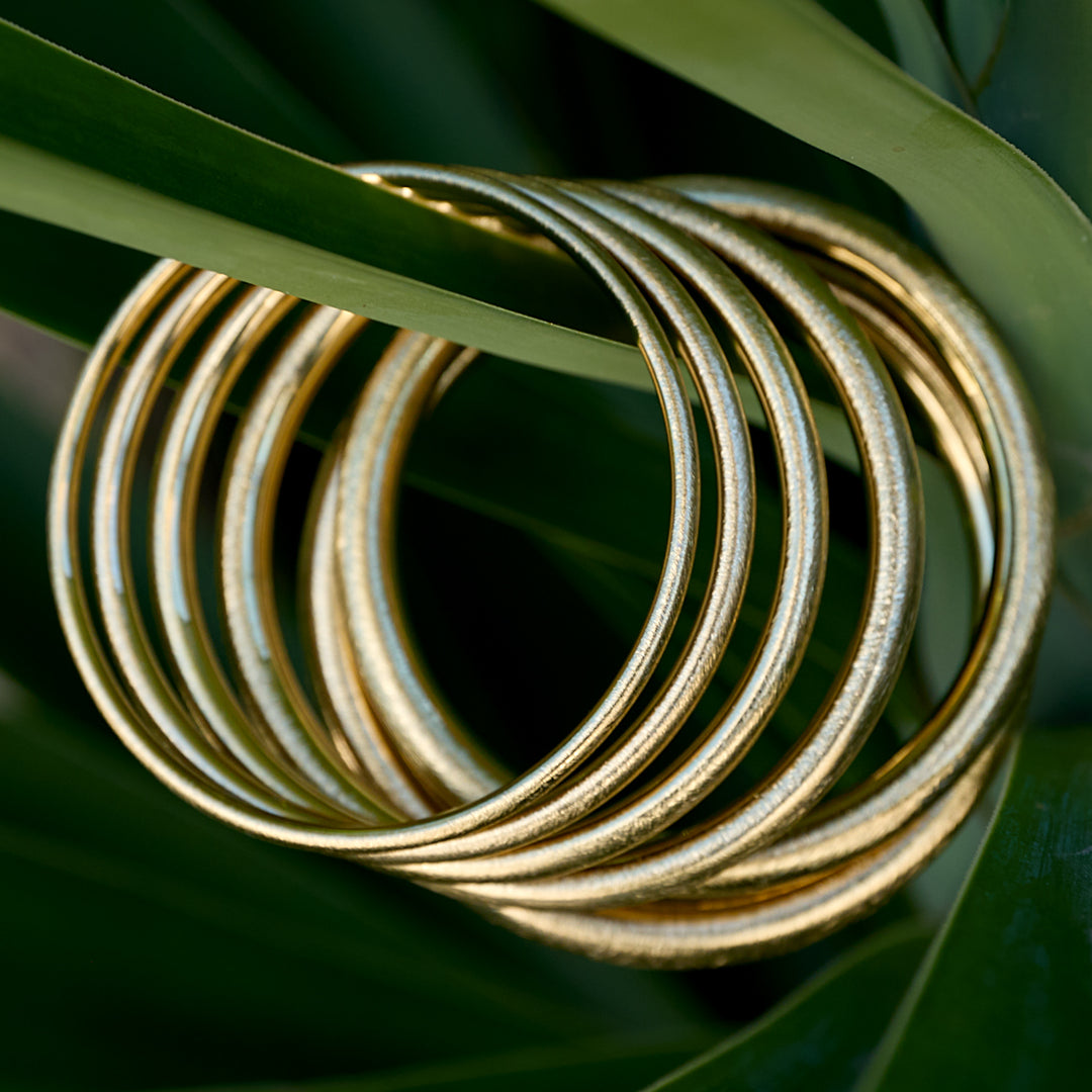 A stack of gold Elara Bangle Bracelets nestled among lush green leaves.