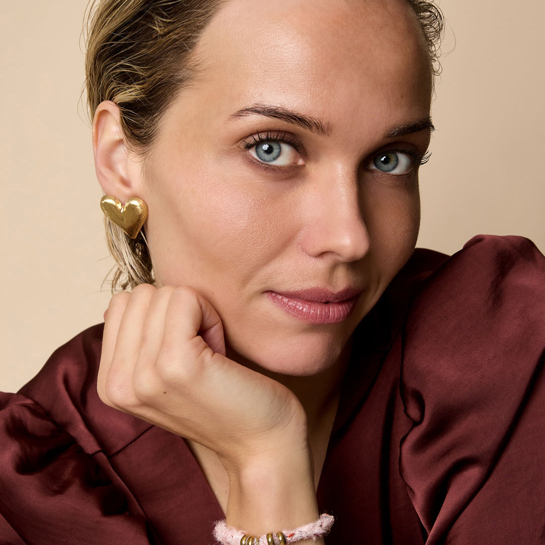 Gold heart stud earrings on a woman's ear, with a matching heart charm bracelet on her wrist.