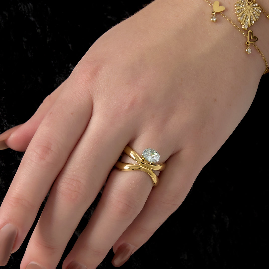 Gold adjustable ring with a clear oval stone on a hand with brown nails and a gold bracelet.
