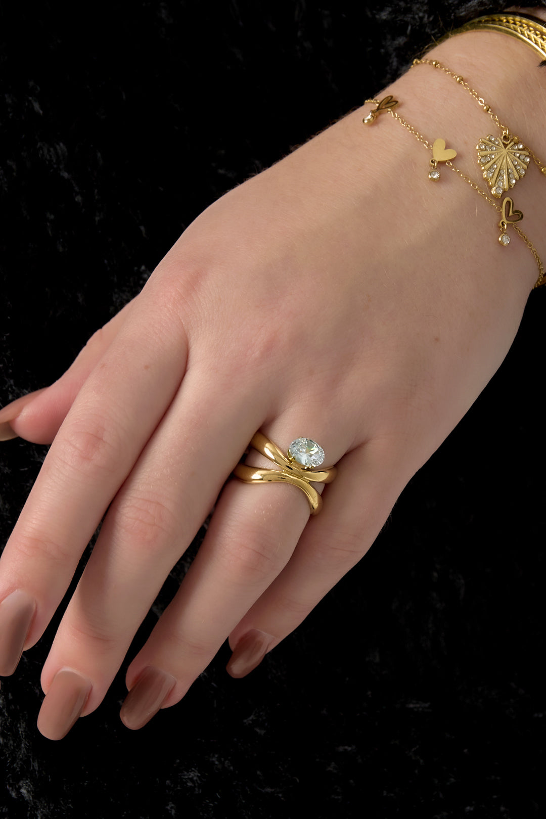 Gold adjustable ring with a clear oval stone on a hand with brown nails and a gold bracelet.