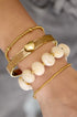 Gold bangle with a heart charm, layered with beaded bracelets and cream-colored beads.