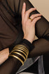 A stack of gold Elara bangle bracelets, 10 mm wide, worn on a wrist with a gold ring.