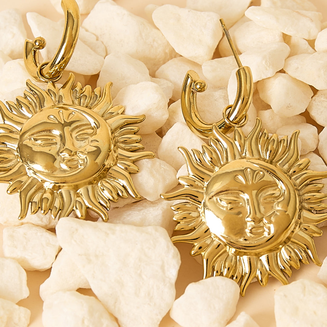 Gold sun drop earrings with detailed faces and rays, resting on white stones.