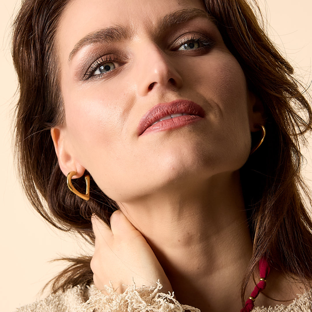 Gold heart hoop earrings worn by a woman with brown hair and blue eyes, wearing a cream textured sweater.