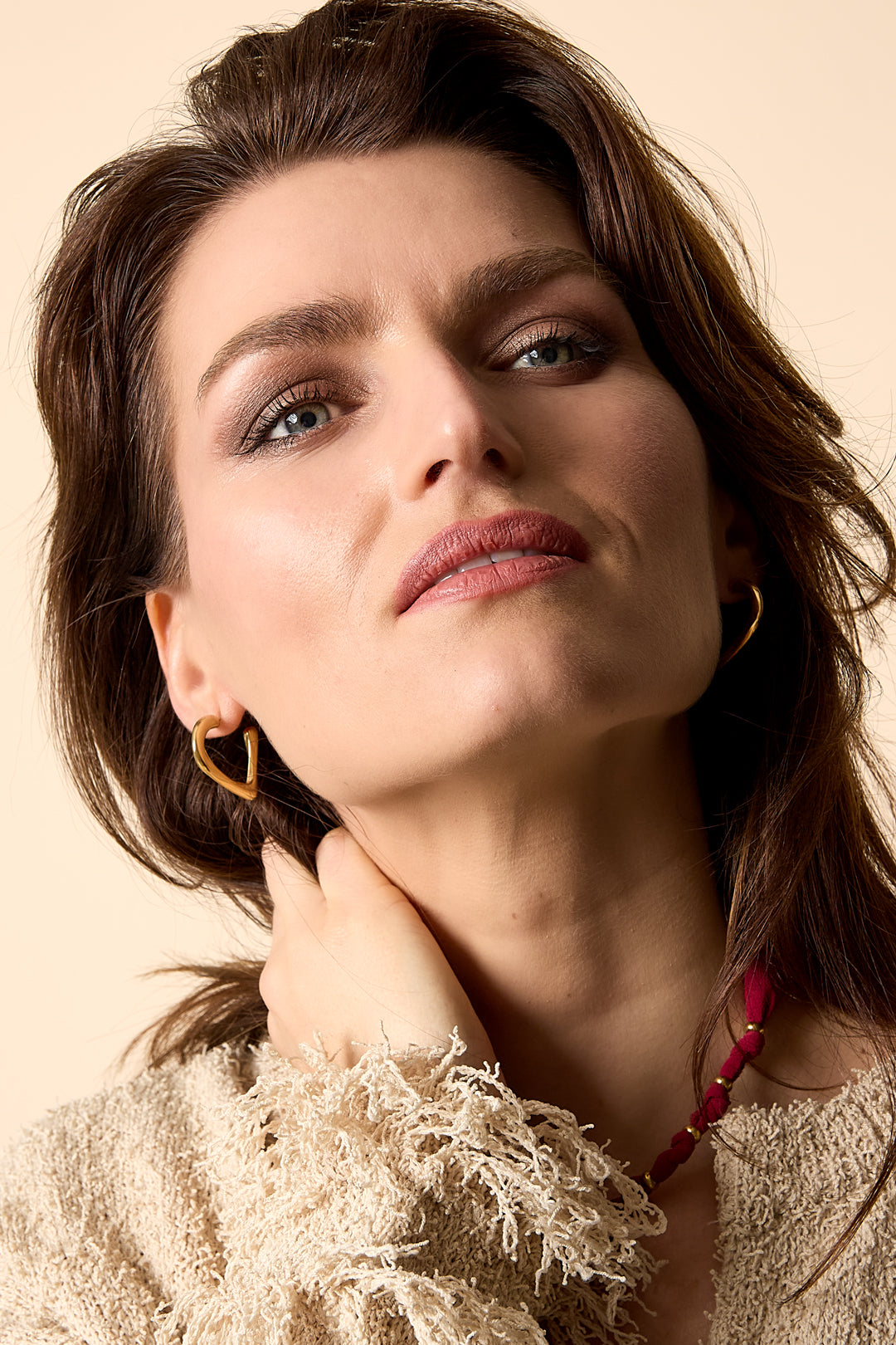 Gold heart hoop earrings worn by a woman with brown hair and blue eyes, wearing a cream textured sweater.