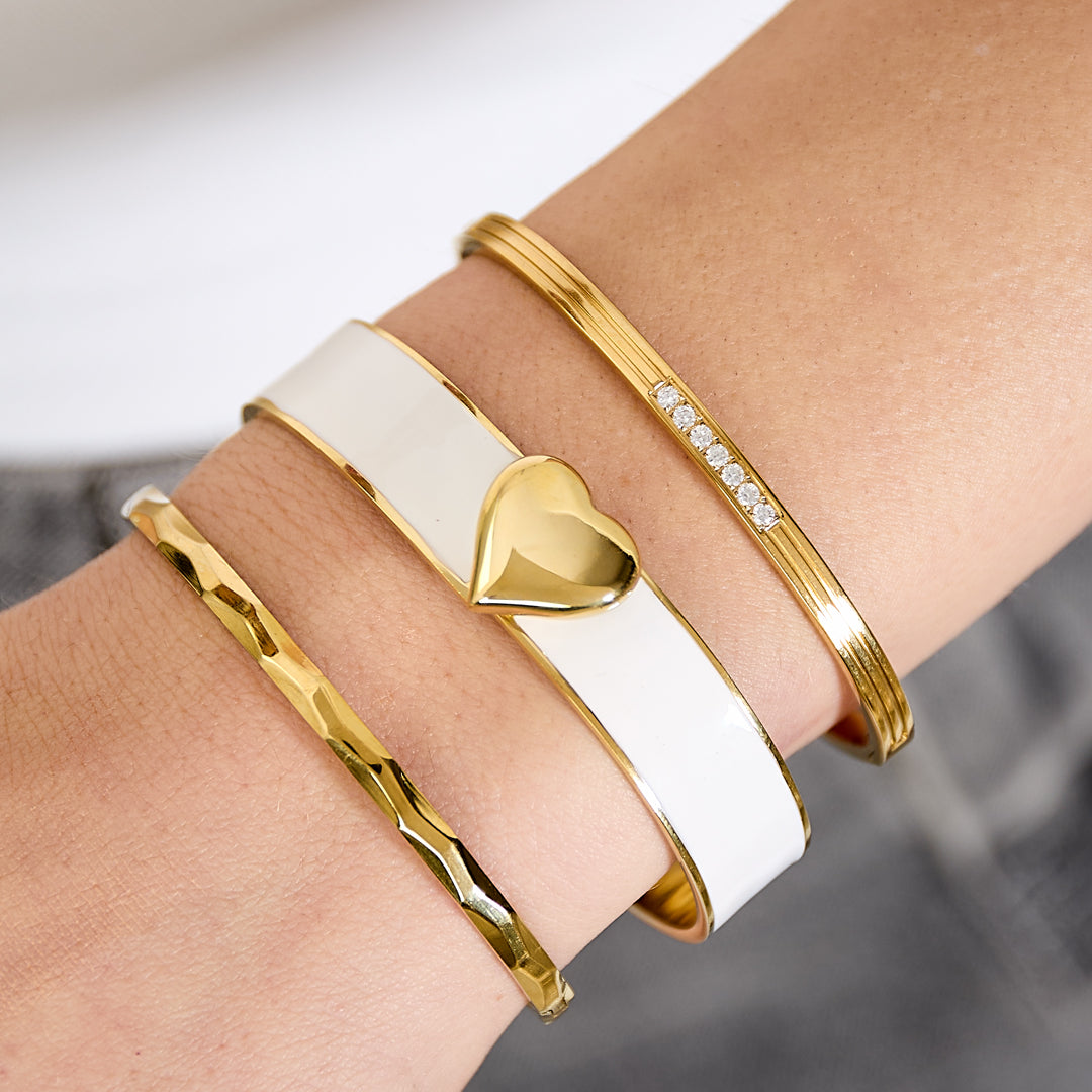 Three gold bangles on a wrist: one with a white band and gold heart, one with small diamonds, and one textured.
