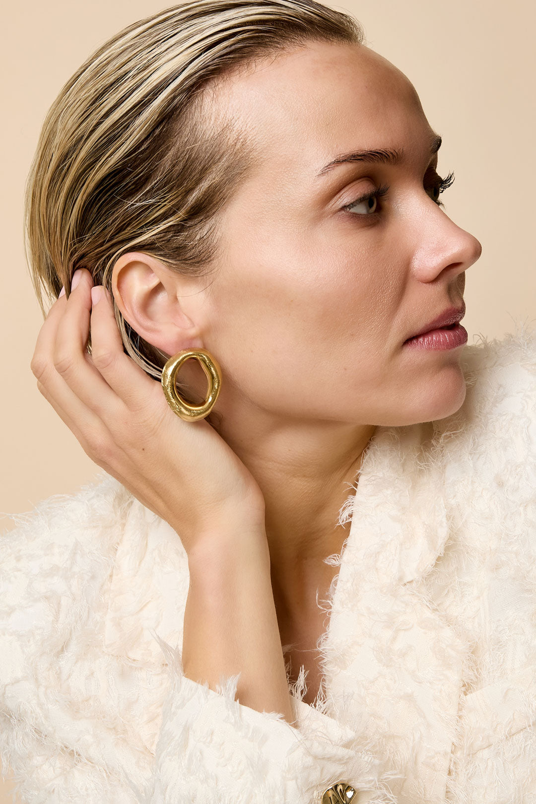 Gold-toned irregular hoop stud earrings on a woman's ear, styled with a textured white jacket.