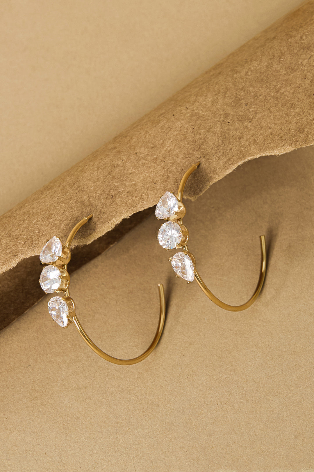 Gold hoop earrings with three clear stones each, set against a brown background.