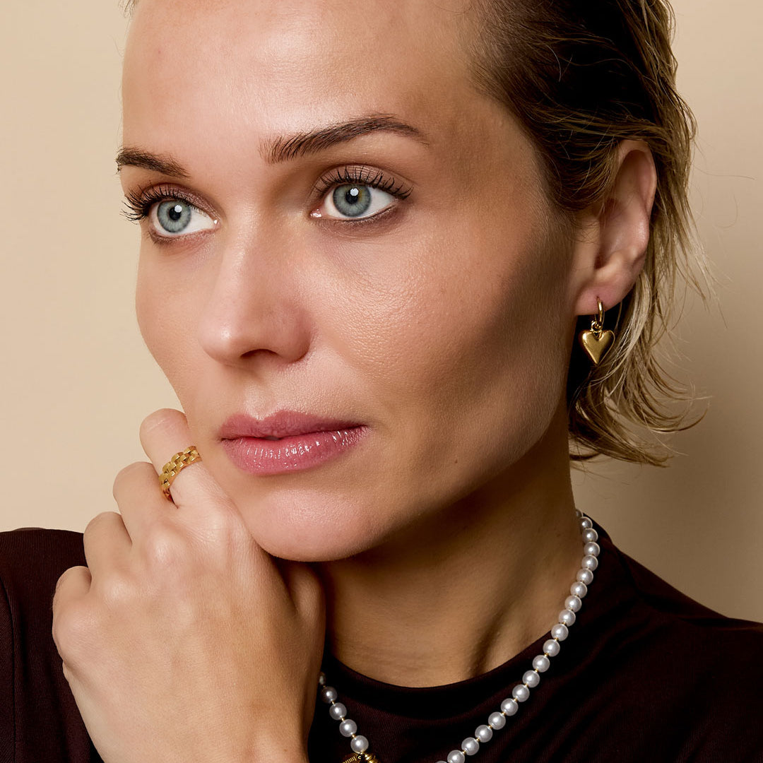 Gold heart drop earrings on a woman with a pearl necklace and gold ring.