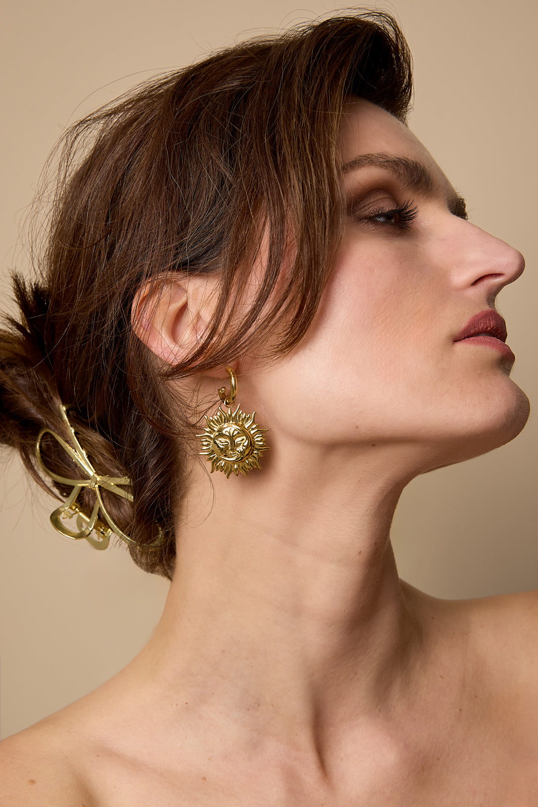 Gold sun drop earrings on a woman's ear, with a gold bow hair clip in her hair.